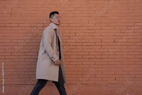 Middle-aged Latino man with glasses, beige trench coat, and brick background poses