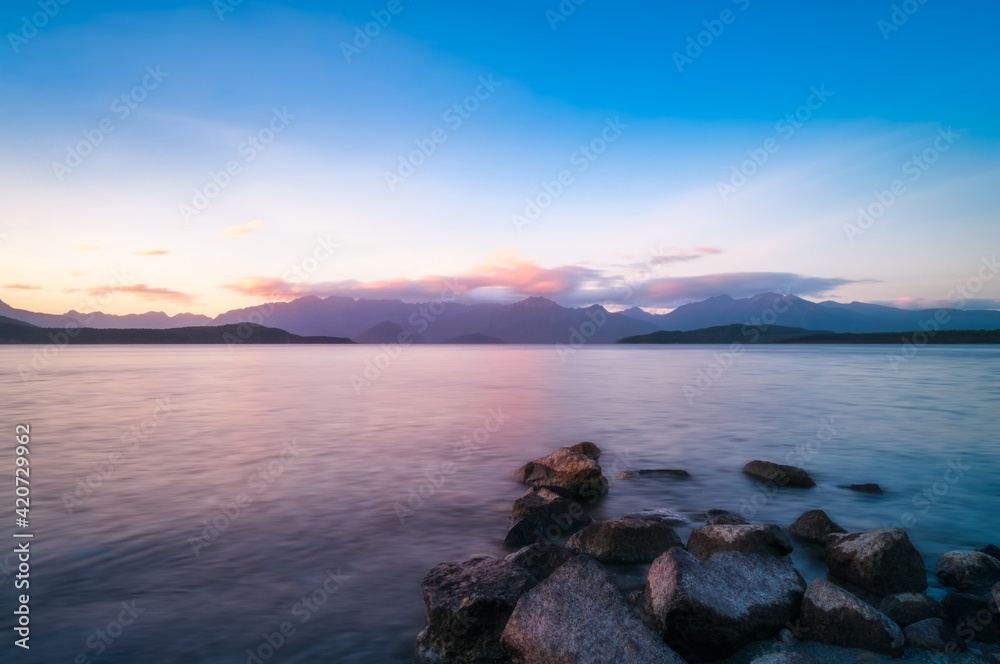 Colorful sunset on the shore at Lake Te Anau with rocks in the water and the mountain range silhouetted in the background in Fiordland National Park, New Zealand, South Island.