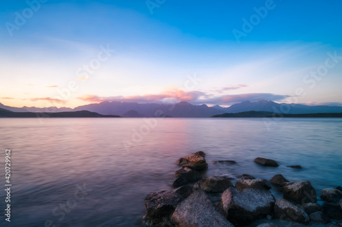 Colorful sunset on the shore at Lake Te Anau with rocks in the water and the mountain range silhouetted in the background in Fiordland National Park, New Zealand, South Island.