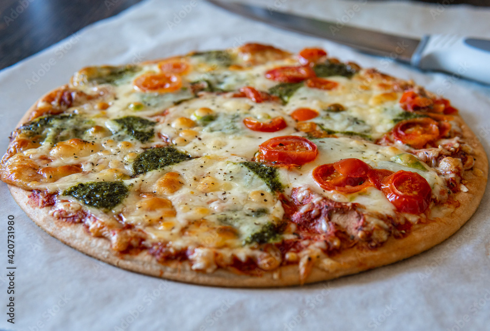 Colorful fresh pizza hot from the oven with mozzarella cheese, pesto sauce and cherry tomatoes. Baked, delicious, gluten free pizza ready to be sliced while placed on baking sheet. Knife in background