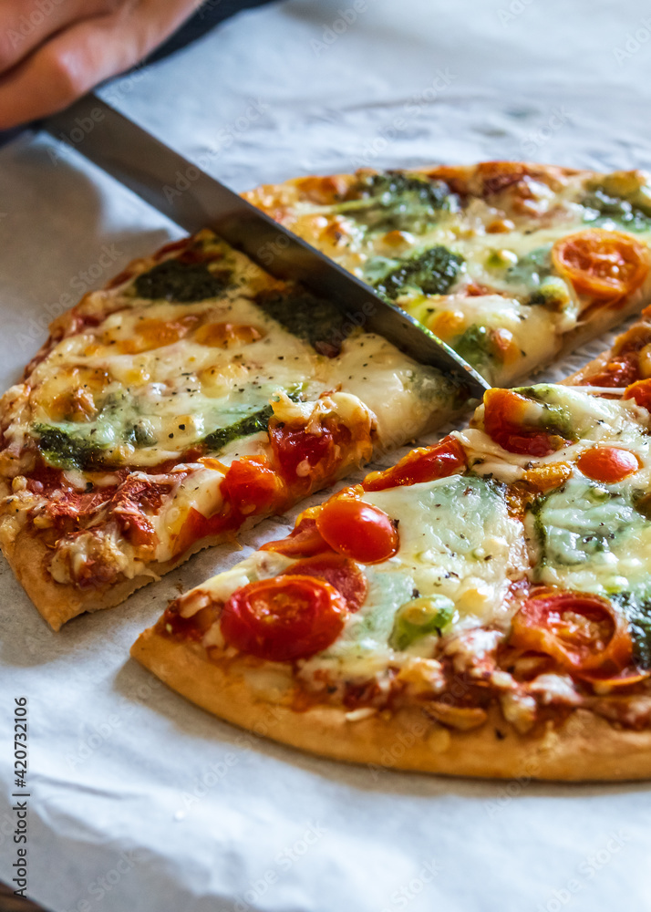 Hand cutting colorful fresh pizza hot from the oven with mozzarella cheese, pesto sauce and cherry tomatoes with a knife. Baked, delicious, gluten free pizza cut into quarters and ready to be served. Hand cutting colorful fresh pizza hot from the oven with mozzarella cheese, pesto sauce and cherry tomatoes with a knife. Baked, delicious, gluten free pizza cut into quarters and ready to be served.
