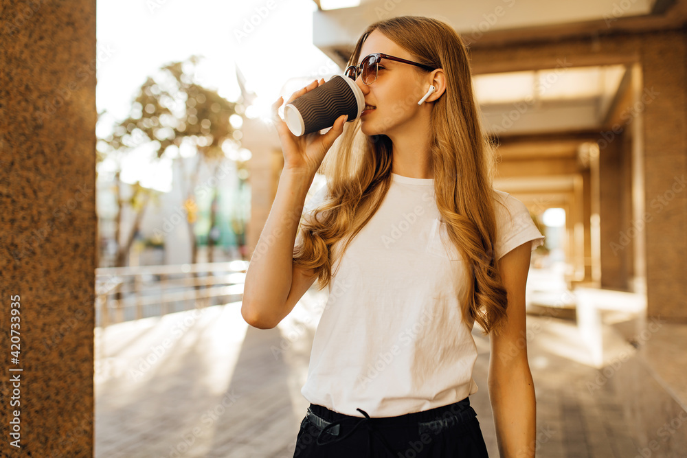 young woman, with wireless headphones, holding coffee cup on city street