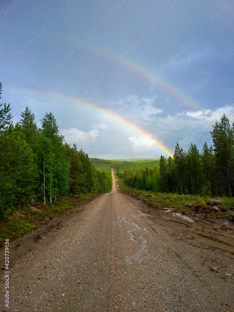 A scenic view of a double rainbow in the cloudy rainy sky over dense Oulanka National Park landscape with a rural path
