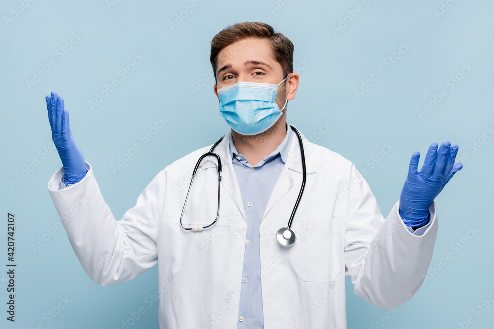 young doctor in medical mask and white coat standing with outstretched hands isolated on blue young doctor in medical mask and white coat standing with outstretched hands isolated on blue
