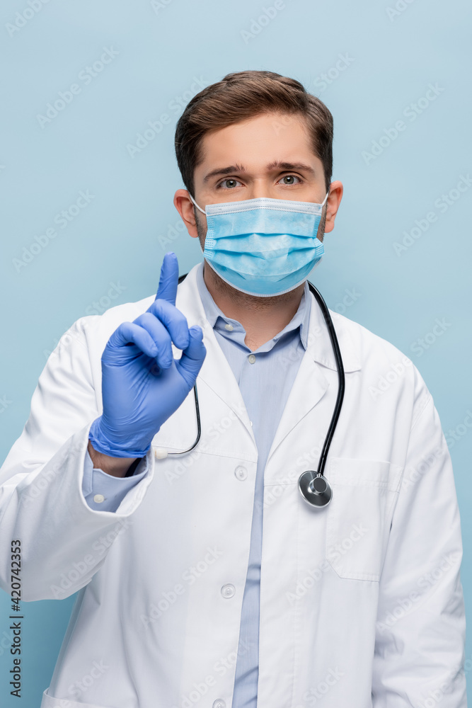 young doctor in medical mask and white coat with stethoscope pointing with finger isolated on blue young doctor in medical mask and white coat with stethoscope pointing with finger isolated on blue