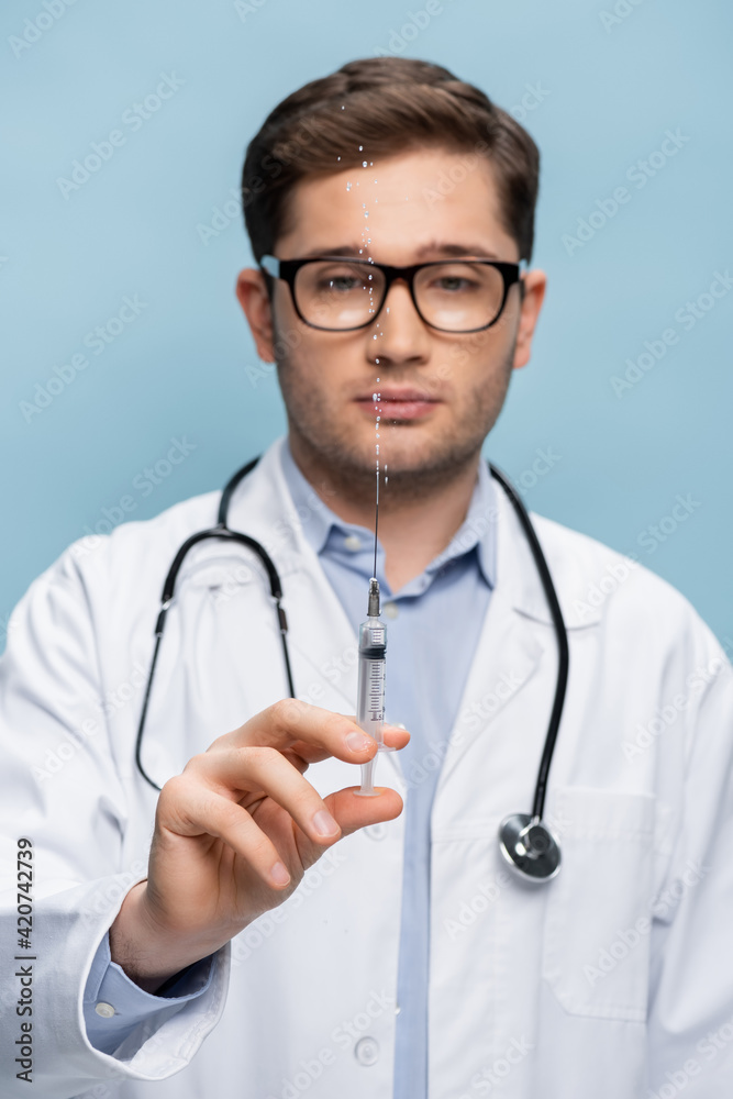 blurred doctor in glasses and white coat holding syringe with vaccine isolated on blue