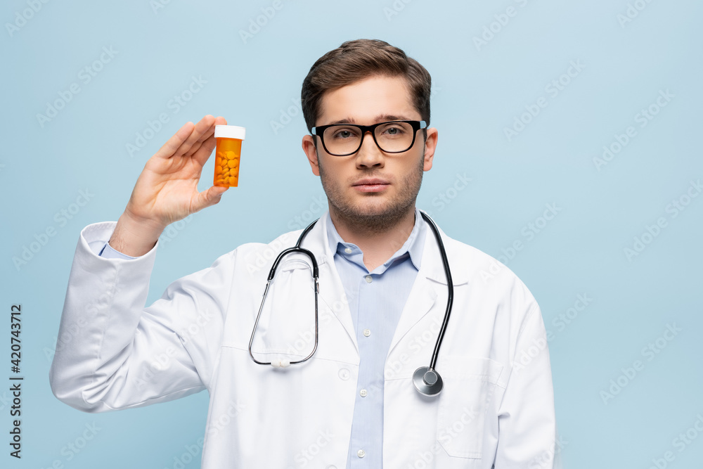 young doctor in glasses and white coat holding bottle with medication isolated on blue young doctor in glasses and white coat holding bottle with medication isolated on blue
