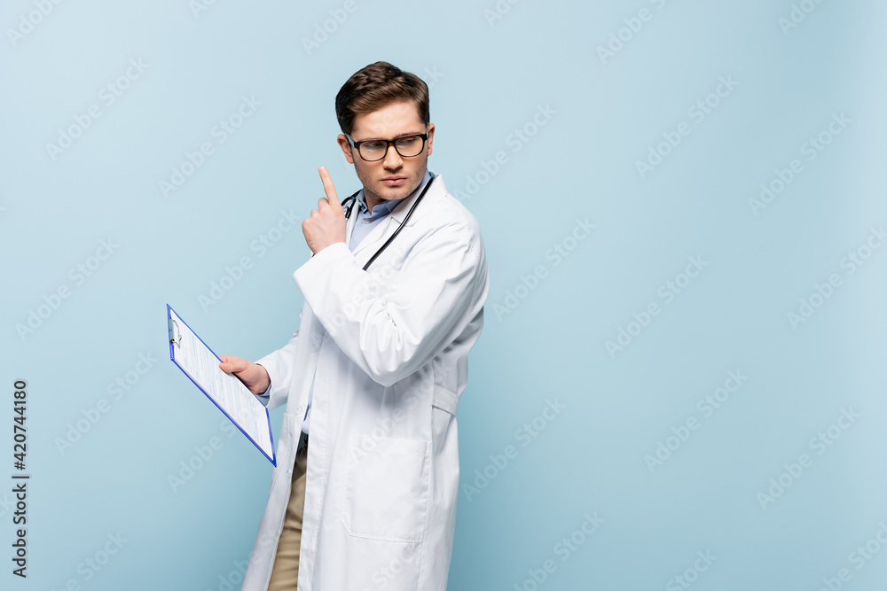 pensive doctor in glasses and white coat holding clipboard and showing idea sign isolated on blue