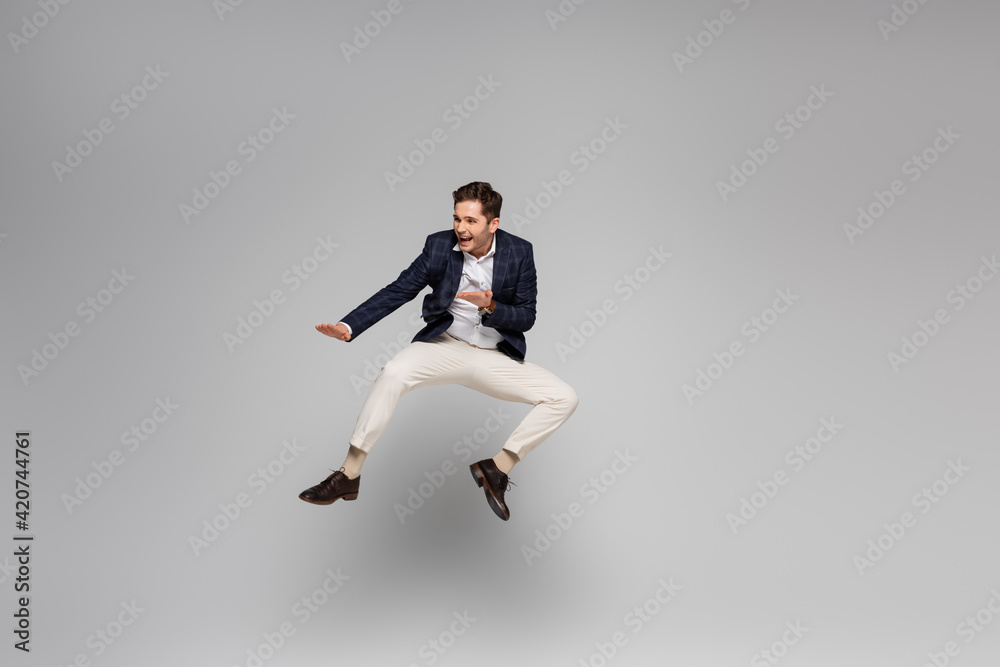 full length of cheerful young man in blazer levitating on grey