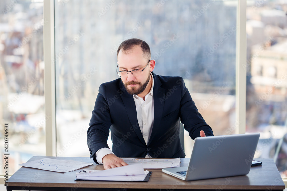 Adult male mentor, director, businessman in glasses and a suit studying documents while sitting at the table. Working day concept Adult male mentor, director, businessman in glasses and a suit studying documents while sitting at the table. Working day concept