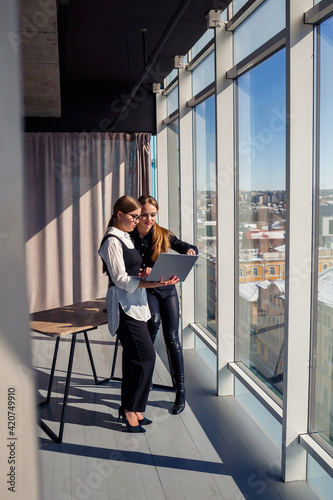 Two successful female architects are talking about a joint project while standing at the window with a laptop. Young women economists dressed in formal attire talking during a break from work