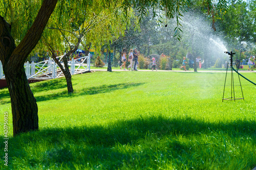 sprinklers water the green lawn in the city park on a summer day, people walking and children playing, bright sunlight and shadows