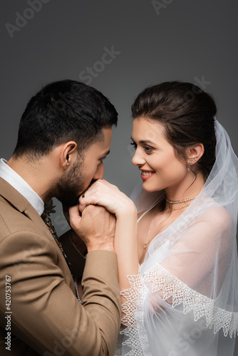 elegant muslim man kissing hands of cheerful bride isolated on grey