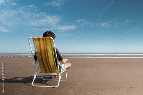 Young man sitting on a deck chair relaxing and enjoying the sunny day at the beach. © Kevin