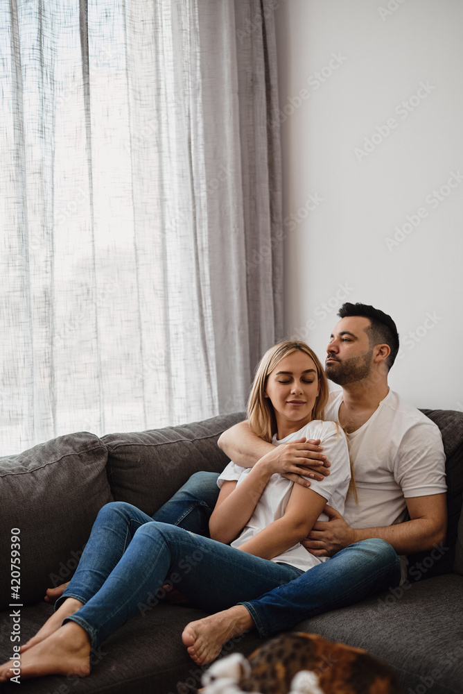 Young woman and man sitting at the couch and hugging each other. Relax at home. Young married couple. Love and romantic relationship