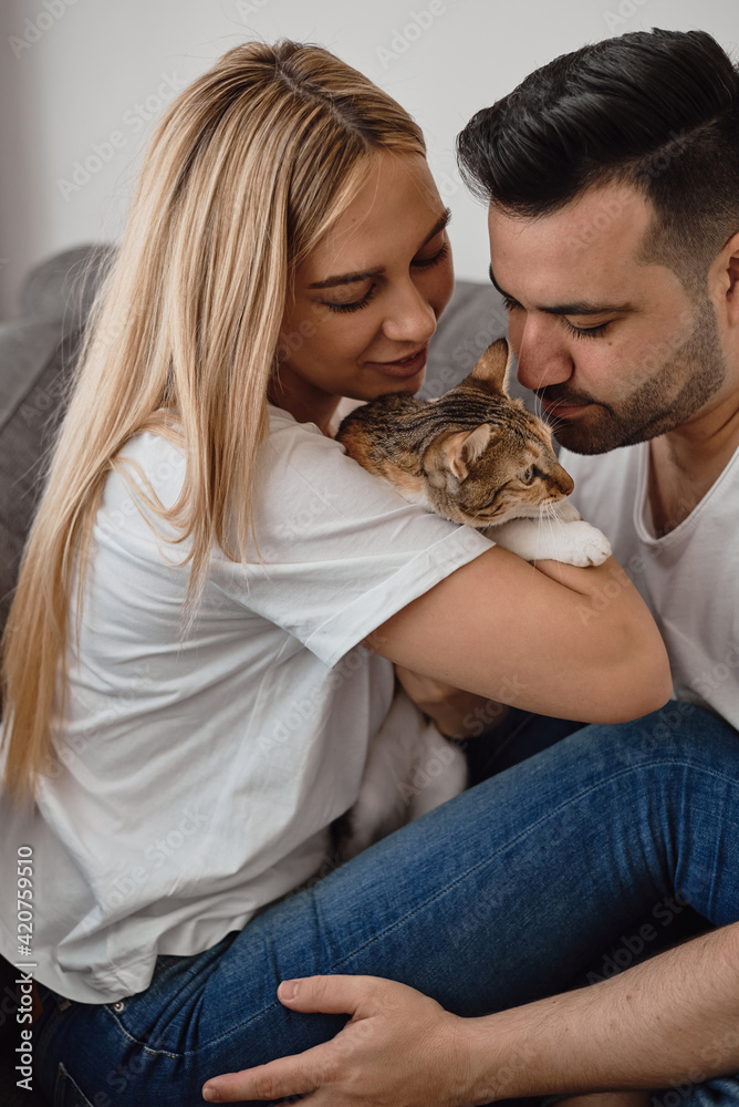 Young couple sitting at sofa and cuddling a small cat. Family, love and romantic relationship. People and pets Young couple sitting at sofa and cuddling a small cat. Family, love and romantic relationship. People and pets