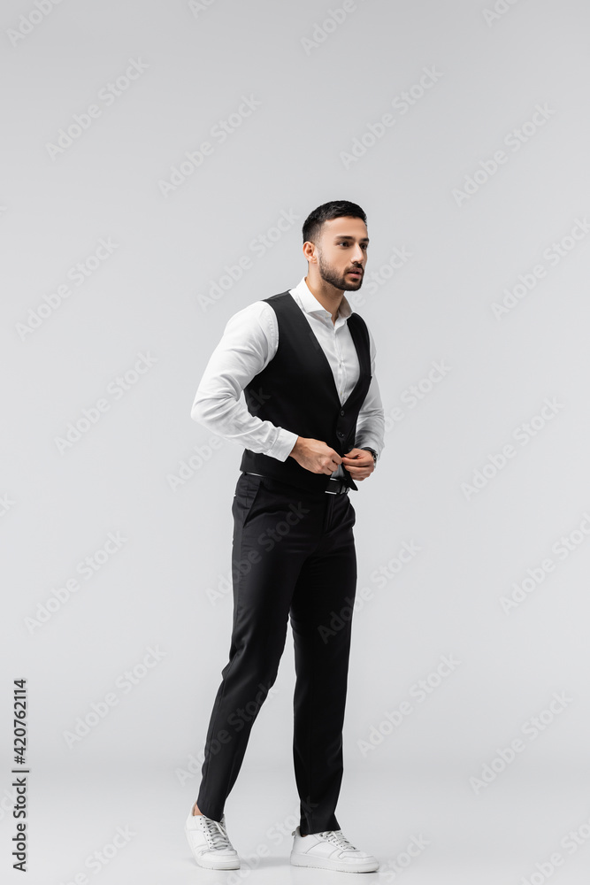 full length view of trendy muslim man buttoning vest while looking away on grey full length view of trendy muslim man buttoning vest while looking away on grey
