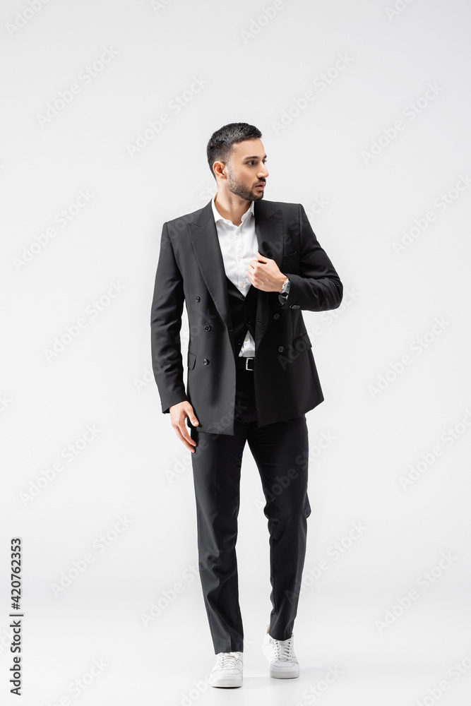full length view of muslim man in black suit and sneakers looking away while walking on grey