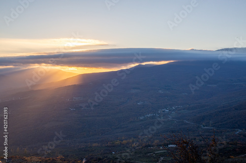 The rays of the setting sun illuminate the mountain valley from under blue clouds