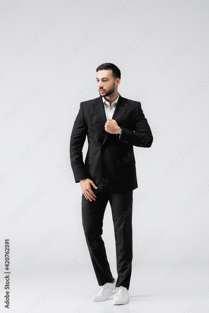young arabian man in black, elegant suit and sneakers looking away while standing on grey young arabian man in black, elegant suit and sneakers looking away while standing on grey