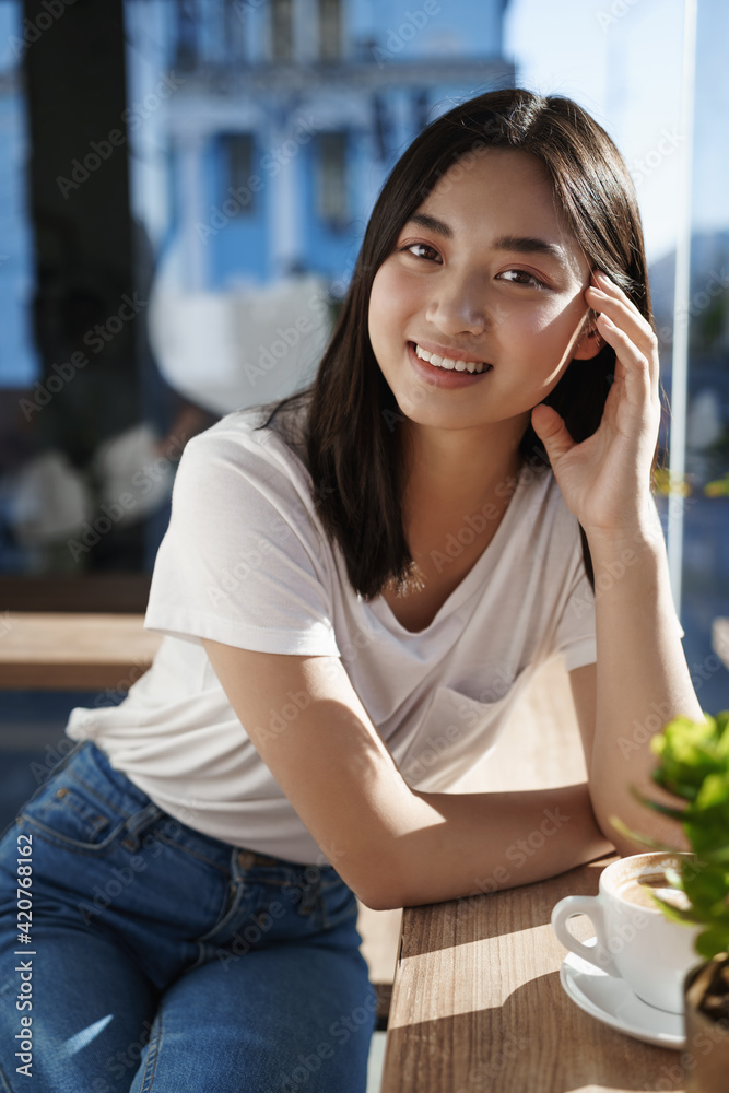 Close up portrait of asian woman sitting in restaurant near window and drinking coffee. Girl in cafe looking happy at camera, smiling