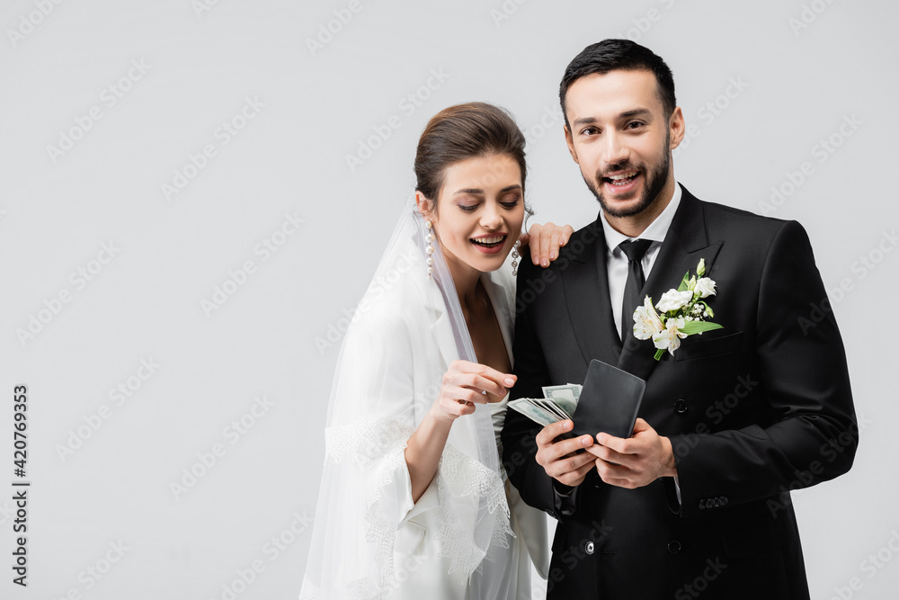 Smiling bride looking at wallet with money in hands of muslim groom isolated on grey Smiling bride looking at wallet with money in hands of muslim groom isolated on grey