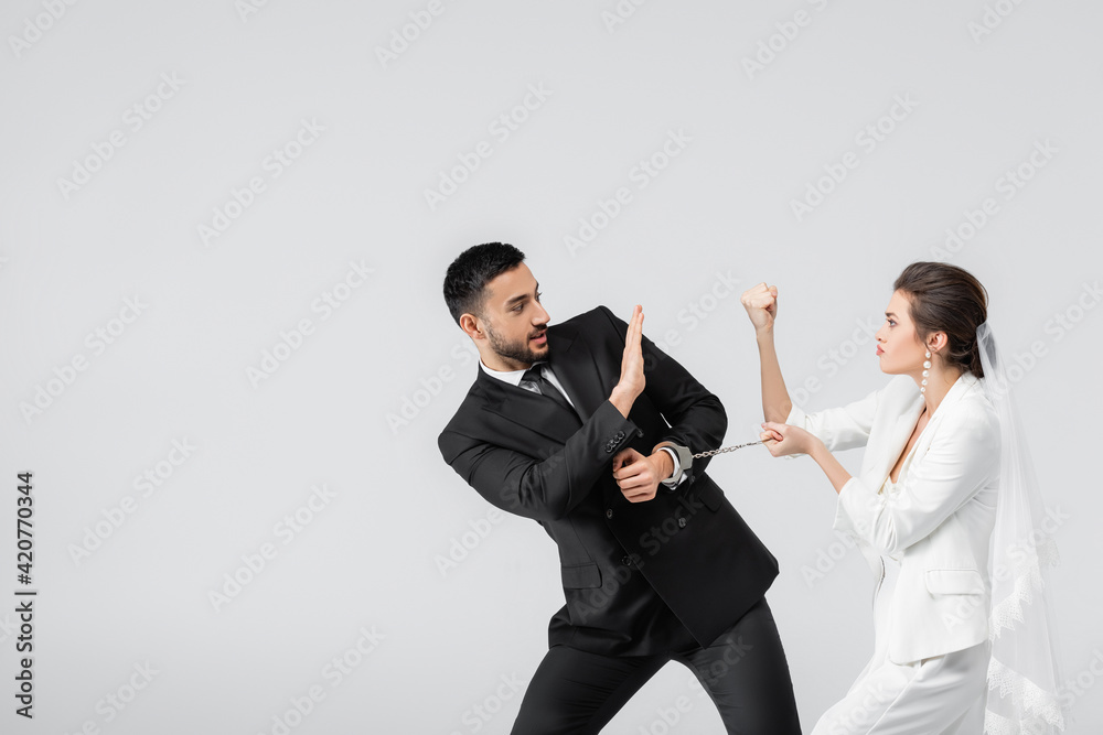 Bride with handcuffs showing fist to muslim groom showing shop gesture isolated on grey Bride with handcuffs showing fist to muslim groom showing shop gesture isolated on grey