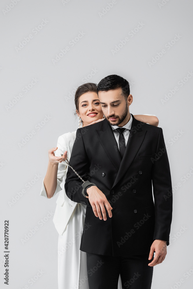 Smiling bride holding handcuffs near muslim boyfriend isolated on grey Smiling bride holding handcuffs near muslim boyfriend isolated on grey