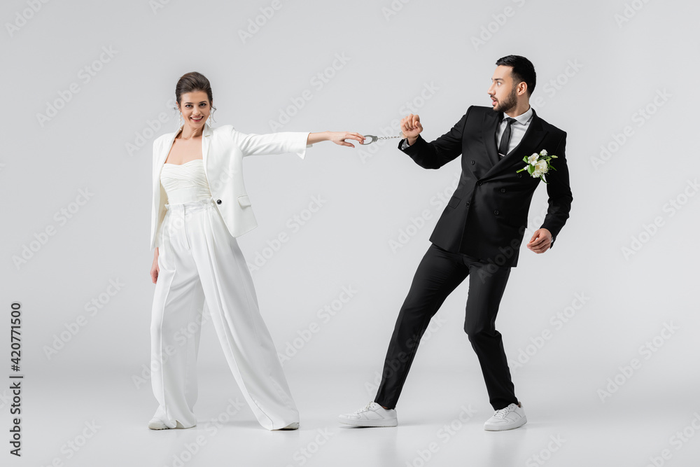 Positive bride pulling handcuffs of muslim groom on white background