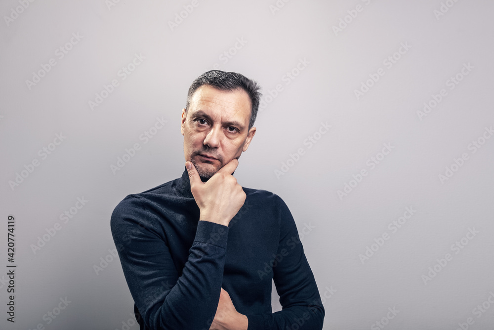 Fototapeta premium A white middle aged man of European appearance with gray in his hair and a slightly unshaven face in a black turtleneck looks proudly at the camera supporting his head by the chin