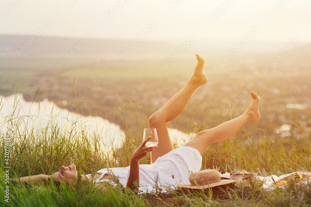 Young beautiful happy woman with white wine in hand is lying on the blanket and enjoying perfect time at summer picnic.