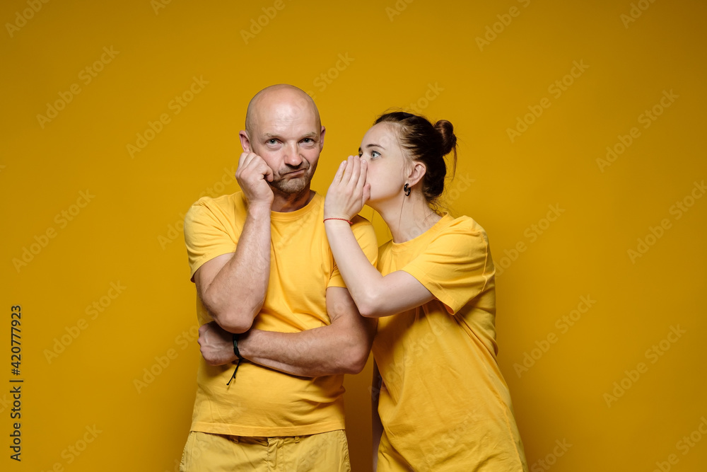 Woman shares a secret or whispers gossip in the ear of a bewildered man who looks thoughtfully into the camera. Yellow background.