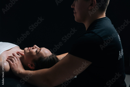 Smiling young woman lying down on massage table during shoulder and neck massage at spa salon. Male masseur professionally massaging shoulders on black background.