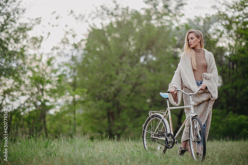 Blonde woman stands among meadow with bicycle.