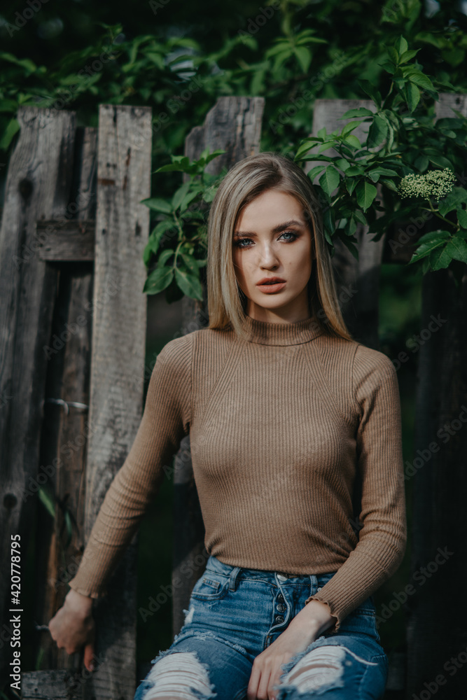 Fototapeta premium Blonde woman sits near wooden fence and elderberry shrub.