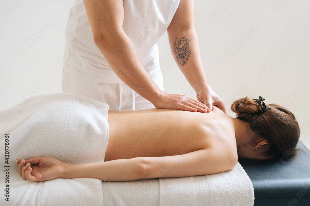 Fototapeta premium Side view of hands of male masseur with strong hands professionally massaging scapulas and shoulders. Beautiful naked young woman with perfect skin getting back massage. 