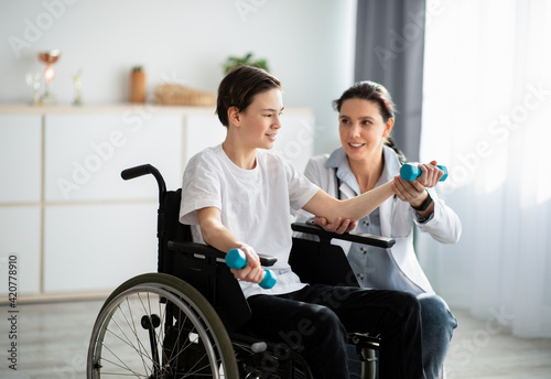 Physical rehabilitation concept. Young physiotherapist helping teenage boy in wheelchair to do exercises at home photo