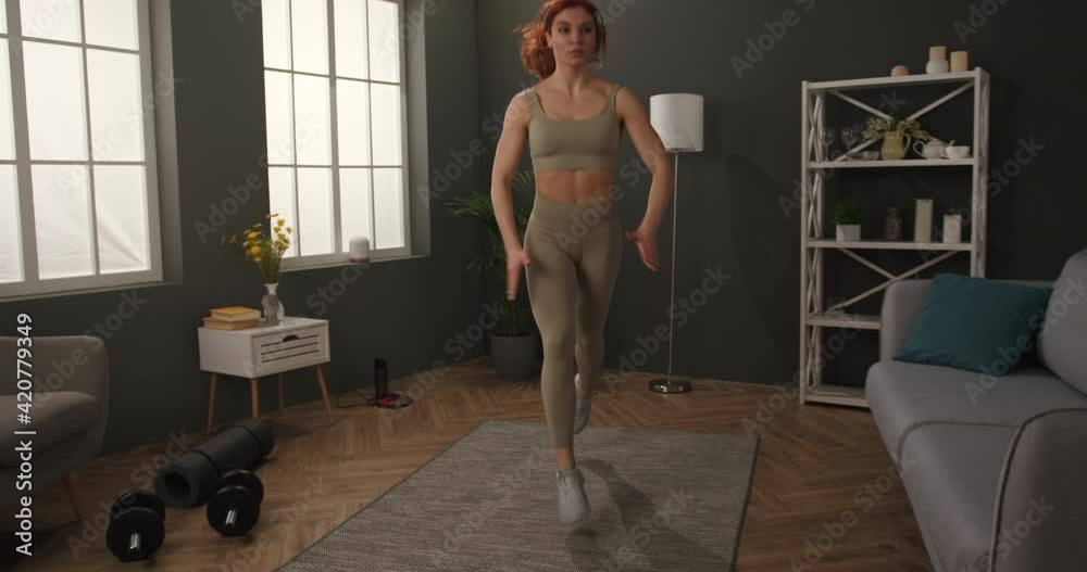 Fit young woman enjoys morning fitness workout at home doing jumping lunges. Casual lady in sportswear training legs.