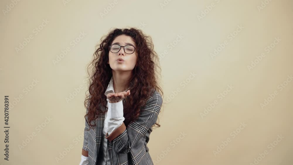 The young woman in love shares kisses with outstretched palm, at the end she smiles with her teeth out. Cretaceous in gray jacket and white shirt, with glasses posing isolated on a beige background in