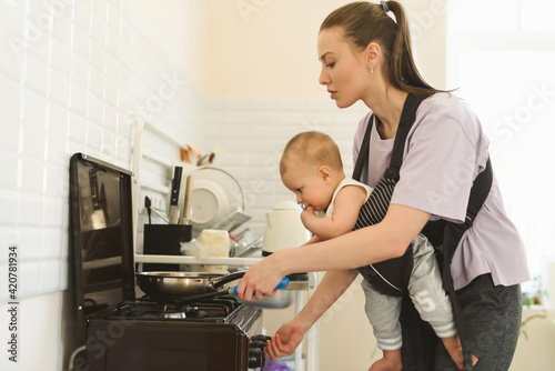 A young mother cooks in the kitchen, the child is fastened to her chest with a baby carrier or babywearing system. The difficulties of motherhood.