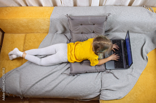 Digital native generation alpha, cute kid lying on yellow sofa with gray blanket and using laptop for chatting and distant learning, online education at home, new normal lifestyle, trendy colors © Maria