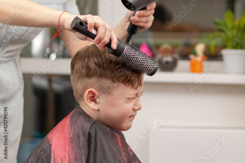 A mother with a hairdryer in her hand in a light blue dress is doing her son's hair at home during the second period of quarantine. selective focus