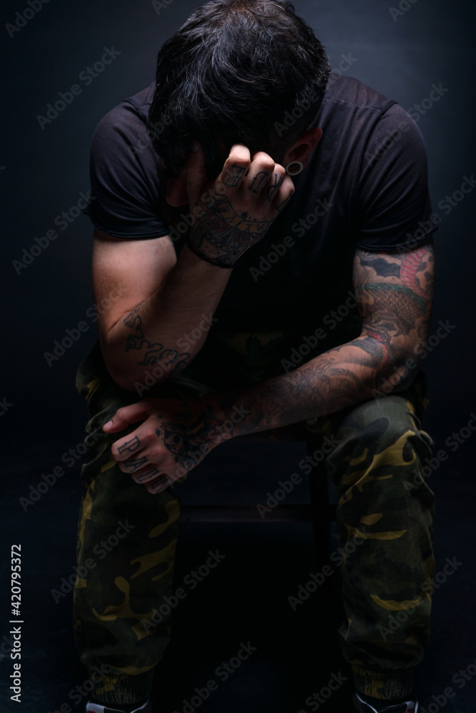 High contrast picture of a bearded male model sitting on a chair and covering his face with his tattooed hands