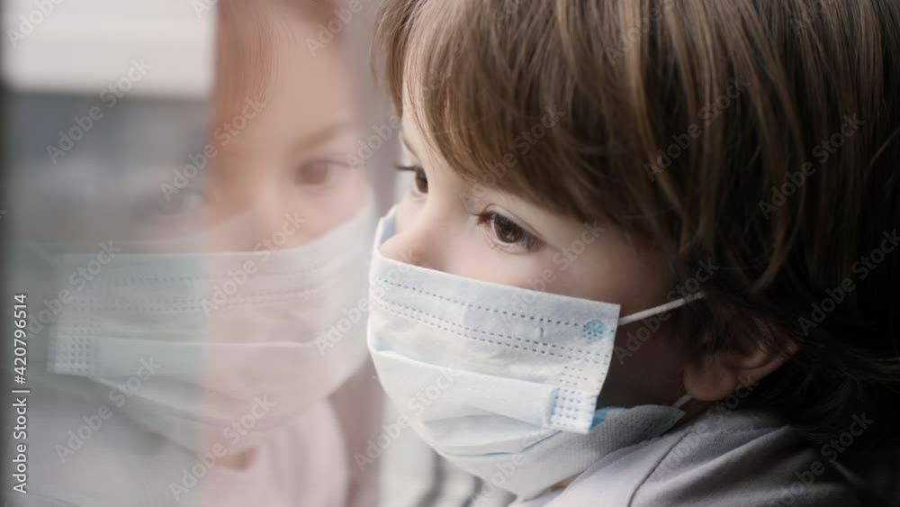 Little Child Boy With Protective Mask, Sad Looking Through Window Worried About Covid-19 Lockdown. Kid In Protection Mask Looking Out Window Home. Quarantine Coronavirus Social Distancing Little Child Boy With Protective Mask, Sad Looking Through Window Worried About Covid-19 Lockdown. Kid In Protection Mask Looking Out Window Home. Quarantine Coronavirus Social Distancing