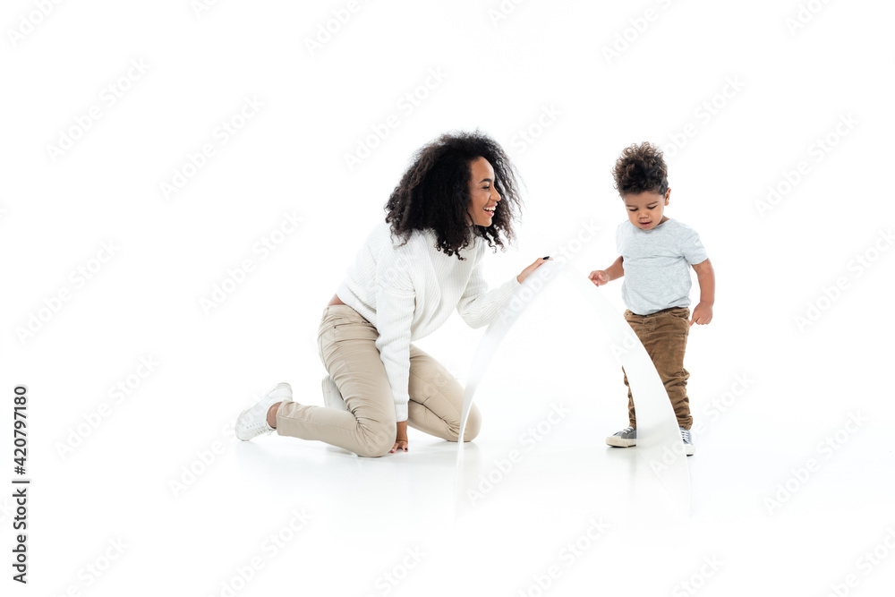 full length view of cheerful african american woman and toddler son near paper roof on white