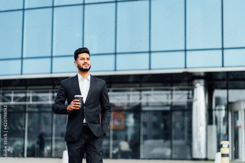 In the photo, an Indian man with coffee in his hands is walking to an important meeting, he is smiling, behind him is a large wooden building, an office worker