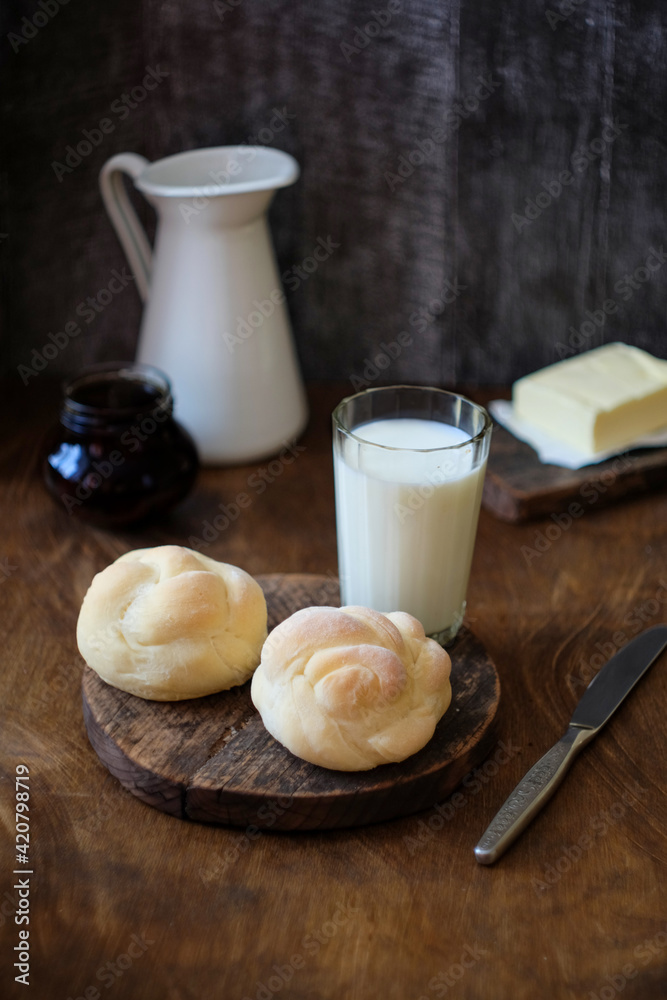 Brioche on a wooden background with a glass of milk, a white jug, butter. Bun. Side view. Brioche on a wooden background with a glass of milk, a white jug, butter. Bun. Side view.