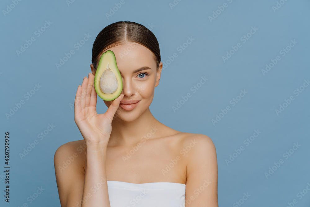 Face care concept. Charming tender woman with combed dark hair has glowing skin covers eye with half of avocado gets essential oils for body treatment wrapped in bath towel isolated on blue wall Face care concept. Charming tender woman with combed dark hair has glowing skin covers eye with half of avocado gets essential oils for body treatment wrapped in bath towel isolated on blue wall