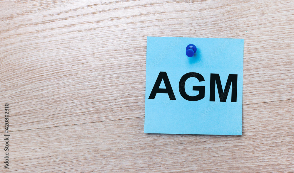 On a light wooden background - a light blue square sticker with the text AGM Annual General Meeting On a light wooden background - a light blue square sticker with the text AGM Annual General Meeting