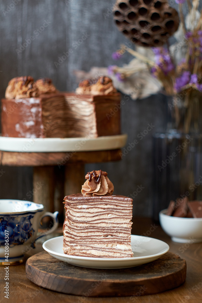 Chocolate crepe cake decorated with cream, chocolate and nuts. Wooden background. In the background is a gray wooden background. Chocolate, coffee, purple flowers. Side view. Chocolate crepe cake decorated with cream, chocolate and nuts. Wooden background. In the background is a gray wooden background. Chocolate, coffee, purple flowers. Side view.